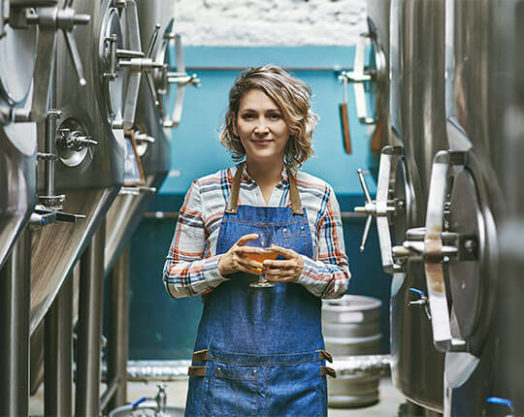 Sonriente mujer cervecera artesanal de pie con un vaso de cerveza extraído directamente de una cuba para su degustación.